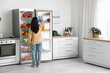 © Pixel-Shot - Young woman taking bottle of water from open fridge full of food in kitchen, back view