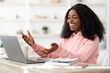 © Prostock-studio - A woman with curly hair smiles and gestures while engaged in a video call. She sits at a desk with a laptop, notebook, and a coffee cup, radiating positivity and engagement.
