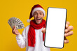 © Prostock-studio - A cheerful man wearing a Santa hat and red scarf is excitedly holding a smartphone in one hand and a stack of cash in the other. The bright yellow background enhances the festive mood.