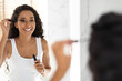 © Prostock-studio - A woman with curly hair smiles in a bright bathroom as she applies mascara in front of a mirror. Sunlight filters through the window, creating a warm atmosphere.