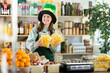 © JackF - Positive teenage girl store clerk in green hat is preparing to meet customers on eve of St. Patricks Day. Employee sells chips, offers snack, wait clients.