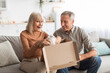 © Prostock-studio - A happy older couple sits on a sofa, joyfully unboxing a surprise package together. The bright living room adds warmth to this special moment.