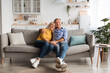 © Prostock-studio - A happy senior couple sits close on a comfortable sofa in a light and airy living room. Warm light fills the space as they smile and share a joyful moment together.