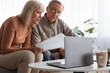 © Prostock-studio - A senior couple sits closely on a couch, looking at documents while having a laptop open on a coffee table. Soft natural light fills the room, creating a cozy atmosphere.