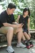 © Masakazu Tokashiki - A man and a woman are sitting on a bench, looking at their cell phones. The man is wearing a black shirt and khaki shorts, while the woman is wearing a black dress. Scene is casual and relaxed