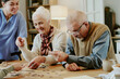 © AnnaStills - Senior Caucasian woman and senior Caucasian man assembling jigsaw puzzle together at table while young adult woman caregiver assisting and smiling in background