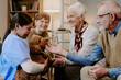 © AnnaStills - Senior Caucasian woman smiling while petting dog held by young adult Hispanic female caregiver, middle aged Caucasian woman and senior Caucasian man sitting nearby interacting with animal