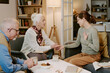 © AnnaStills - Senior Caucasian woman holding hand of middle aged Caucasian woman while sitting on sofa, senior Caucasian man observing conversation, three people interacting in living room