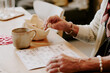 © AnnaStills - Senior Caucasian woman sitting at table playing bingo, holding numbered bingo chip in hand, marking bingo card, wearing bracelet and sweater, coffee mug on table