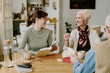© AnnaStills - Senior Caucasian woman smiling and gesturing while sitting at table with middle aged Caucasian woman holding book and middle aged Caucasian man, engaging in conversation