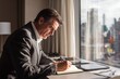 © Alona - Mature businessman writing notes at a hotel room desk, morning light, focused and professional