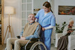 © AnnaStills - Caucasian senior man with disability sitting in wheelchair being assisted by young adult Hispanic woman caregiver while elderly Caucasian woman reading book in background