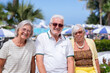 © luciano - Three cheerful senior friends enjoy a sunny day at a seaside resort in a vibrant beach atmosphere with umbrellas, palm trees, and blue sky, expressing relaxation, friendship, and vacation happiness