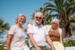 © luciano - Three cheerful senior friends enjoy a sunny day at a seaside resort in a vibrant beach atmosphere with umbrellas, palm trees, and blue sky, expressing relaxation, friendship, and vacation happiness