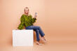 © deagreez - Beautiful mature woman with blonde hair sits on a white block holding a smartphone wearing a green ruffle blouse and jeans against a beige background