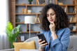 © Liubomir - Young woman with curly hair sitting on a couch at home, focusing on a smartphone screen with a concerned expression, contemplating a difficult message or shocking news