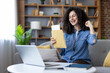 © Liubomir - Happy young woman celebrating exciting news, opening an acceptance letter with a fist pump, sitting on a sofa with a laptop at home or in a student dormitory