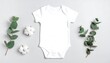 © Rony - A white baby onesie sits centered on a light gray surface, flanked by eucalyptus sprigs and fluffy cotton blooms