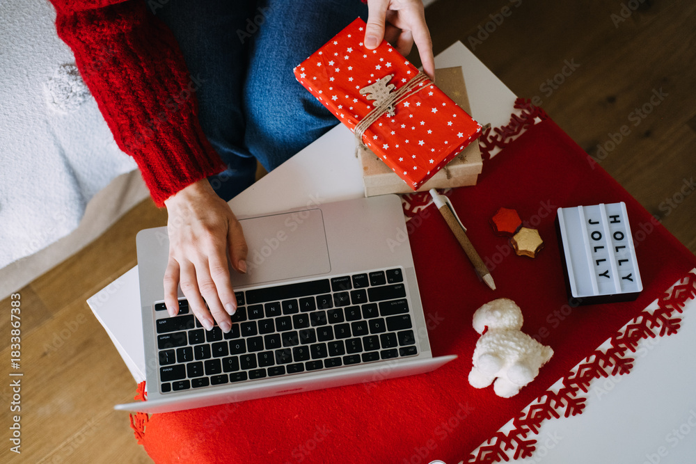 Person writes CHRISTMAS SHOPPING LIST in notebook while holding small gift box on white coffee table. Online shopping, seasonal ecommerce, Christmas sales, holiday planning.