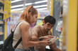 © Chocheng channel - A young couple walks hand-in-hand down a brightly lit retail aisle with high shelves full of colorful products. The woman is pointing or gesturing towards the products, indicating a selection.