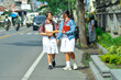 © agratitudesign - Two cheerful teenage girls in school uniforms carrying notebooks walk side-by-side on a paved sidewalk beside a busy road with traffic and traditional stone buildings.