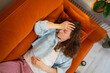 © wifesun - Young woman lying on an orange sofa, struggling with weakness, nausea, stomach pain, and a headache, placing a hand on her stomach and forehead, suffering from illness, sickness, or toxicosis