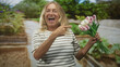© Krakenimages.com - Blonde senior woman holding pink tulips points finger and places hand over chest in a garden with raised beds; gentle gratitude.