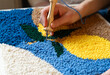 © Natali - Person using a hook to create a design on a colorful rug at home during daytime