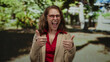 © Krakenimages.com - Young woman with red glasses expressing excitement and positivity with thumbs up in a sunny park setting surrounded by lush green backdrop implying outdoor leisure.