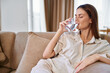 © SHOTPRIME STUDIO - Woman drinking water from a glass on a sofa at home, relaxed portrait of a healthy adult resting in casual clothes, hydration, comfort and calm lifestyle morning moment.