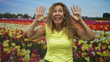 © Krakenimages.com - Woman in yellow sleeveless top raises both hands near face with palms spread in studio tulip field backdrop, smiling with wide eyes and open mouth; playful surprise.