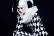 © cmapuk_0nline - A sad Pierrot or jester in a black and white harlequin costume with a ruff collar, crying a single black tear. Dramatic portrait on a dark background.