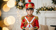 © NeuroCake - Portrait of a cute boy dressed as a nutcracker soldier holding a Christmas gift. Child in red uniform with present box. Festive holiday background with fireplace