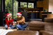 © Wavebreak Media - Diverse couple wearing Santa hats sitting in living room wrapping presents with ribbon, copy space