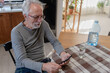 © lordn - Elderly man sitting at a kitchen table, focused on his smartphone, with natural light illuminating the cozy modern kitchen behind him