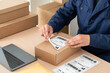 © cunaplus - Latin man preparing a package for shipping, sticking priority mail label on a cardboard box at his startup business office