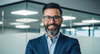 © abu - Close up of a smiling, bearded man wearing glasses and a blue suit jacket. He stands confidently in a modern office with a blurred background. Professional and successful.