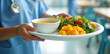 © Maryna - Healthcare worker in blue uniform brings tray with soup salad and fruit to patient. Hospital dining service offers healthy meal for wellness recovery. Nutrition care supports healing.