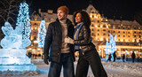 Happy interracial couple enjoying a winter night in a festive city square. Romantic man and woman standing near Christmas lights and ice sculpture