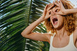 © SHOTPRIME STUDIO - young woman with curly hair playfully making glasses gesture near green tropical palm leaves in bright sunlight, expressing fun and carefree summer vibes