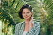 © SHOTPRIME STUDIO - Portrait of a smiling young woman standing outdoors among tropical plants, wearing a green plaid shirt and natural makeup.