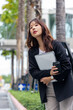 © sorapop - Young Asian woman in business suit on busy street for work, carrying laptop and coffee, observing surroundings in vibrant city environment, looking focused and determined