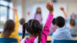 © Curioso.Photography - Children raising their hands in a classroom during a lesson with the teacher visible at the front.