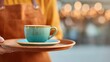 © Maryna - Person is holding a tray with a blue coffee cup on it. The tray is brown and the person is wearing an apron. The scene is set in a restaurant or cafe, and the person is likely a server or a barista