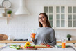 © BestForYou - Woman in gray sweater is holding glass of orange juice and smiling at the camera in kitchen with white brick
