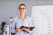 © Prostock-studio - A cheerful teacher stands in front of a whiteboard with French grammar notes. She holds a notebook and smiles, encouraging students as a flag adds color to the setting.