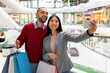 © Prostock-studio - A young couple smiles as they pose for a selfie at a bustling shopping mall. They hold colorful shopping bags, capturing a joyful moment together.