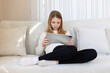 © Iryna - Teen girl sitting on sofa using tablet. Young girl sitting on a bright sofa, focused on a digital tablet in a cozy, sunlit modern living room.