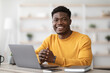 © Prostock-studio - Portrait of positive young black guy in yellow sweater sitting at table, working on laptop, sending emails, office interior. Cheerful african american man freelancer working from home, copy space