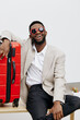 © SHOTPRIME STUDIO - Smiling man with dark skin wearing heart-shaped sunglasses and beige blazer, sitting relaxed with luggage on a white background, casual lifestyle portrait. Happiness, travel, vacation, joy, leisure